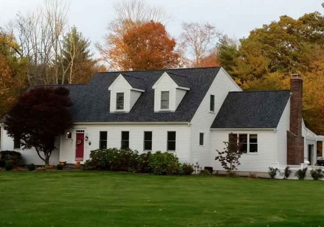 This is the image of a two-stories white house with windows with gables with numerous roof peaks with a black shingles adorning the roof. The house is one of many homes in the South Shore towns of Duxbury, Marshfield, Pembroke, Quincy, Hanover, Norwell, Plymouth, Weymouth, Cohasset, and Scituate that our South Shore roofing contractors' business has done roof repair, roof replacement, roof installation, etc. work on.