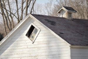 This is the image of about the top third of an old white house. the house looks neglected as paint looks old and deteriorating and the roof of the house, that has a small turret/mini roof looking thing on it, is deteriorating as well. Some shingles are missing from various spots on the piece of roof that is shown. This image is shown to support our South Shore roofing company's blog post regarding how to know when it's time to replace your roof?