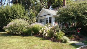 This image shows a modest white home surrounded in front by various flowers and blooming plants.